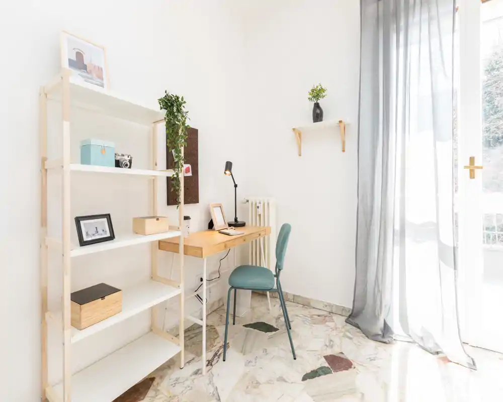 Área de estar luminosa y minimalista con un escritorio de madera como rincón de trabajo, estantería y puerta balcón con cortina translúcida.