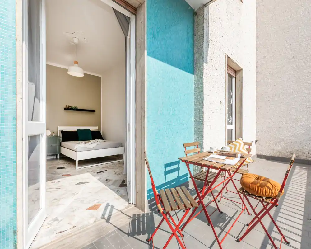 Dormitorio soleado que se abre a una terraza con baldosas y una pequeña mesa tipo bistró. La habitación tiene una cama blanca moderna, mesita y cojines, con mucha luz natural por la puerta abierta.