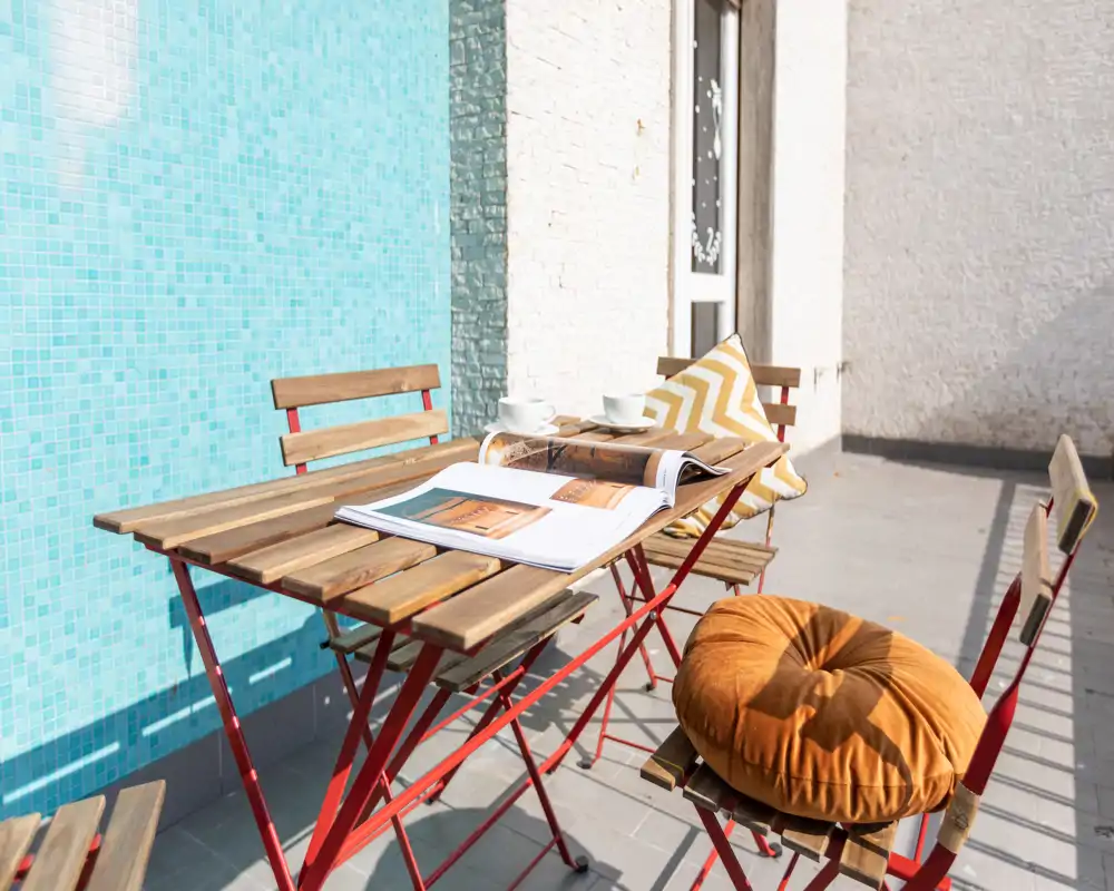 Terraza soleada con mesa bistró de madera y sillas plegables, ambientada con cojines y una revista abierta — perfecta para tomar un café por la mañana o leer al aire libre.