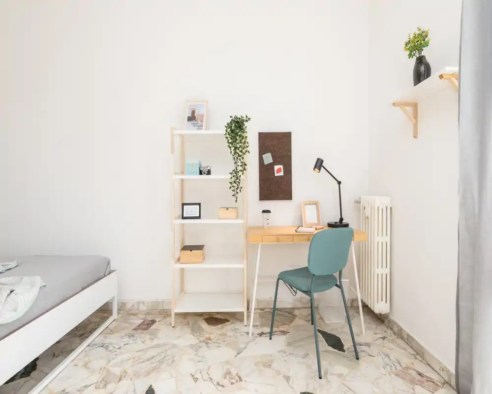 Rincón de dormitorio luminoso y minimalista con cama parcialmente visible, estantería y escritorio ordenado, ideal para trabajar desde casa. Paleta neutra y plantas que crean un ambiente tranquilo y acogedor.