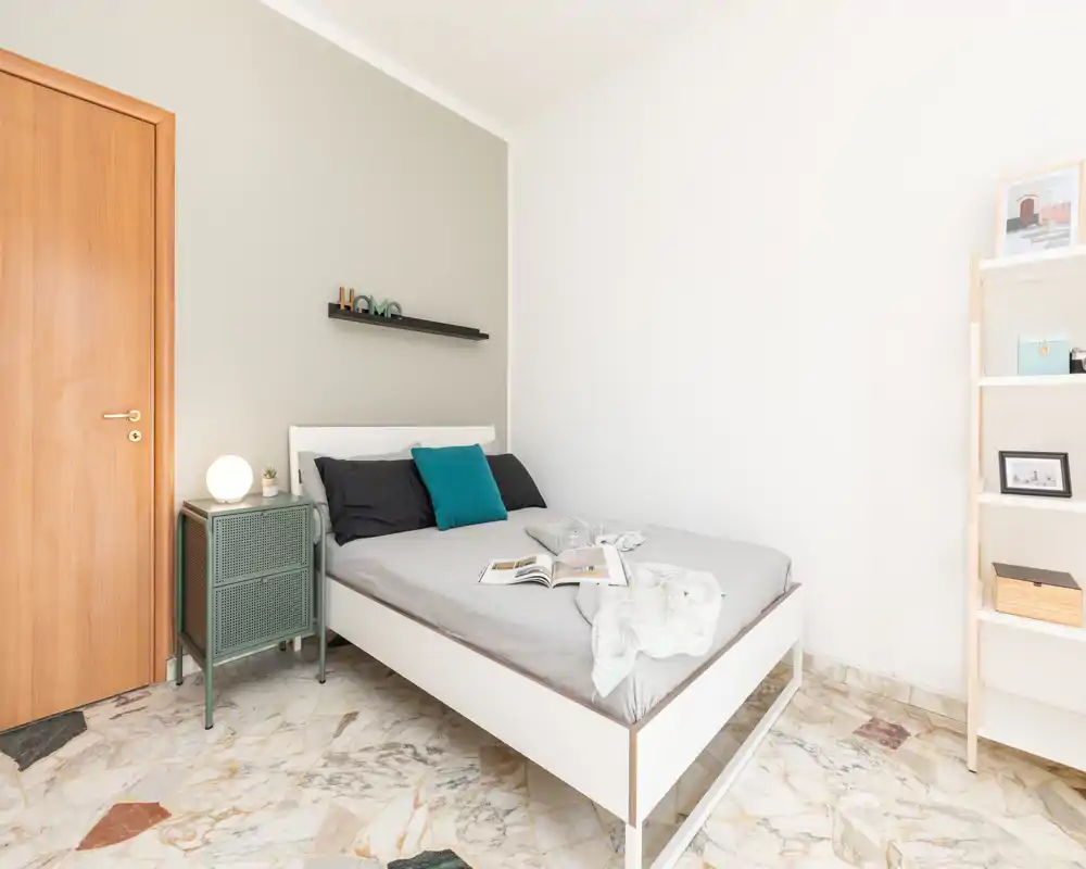 Dormitorio luminoso y minimalista con cama doble bien hecha, mesita y estantería abierta; tonos neutros y suelo de mármol que crean un ambiente moderno y relajado.