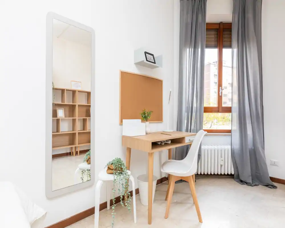 Rincón de dormitorio luminoso y minimalista con escritorio de madera clara, silla y ventana; una pequeña porción de la cama se ve en el borde izquierdo. El espacio es limpio y aireado, ideal para un alquiler compacto o zona de trabajo.