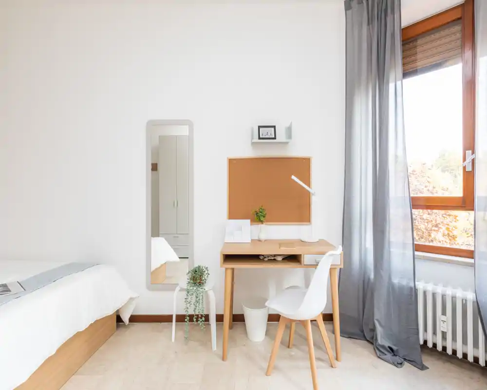 Dormitorio luminoso y minimalista con estructura de cama de madera, pequeño escritorio y luz natural desde la ventana. Paleta neutra y decoración sencilla que crean un ambiente tranquilo y ventilado.