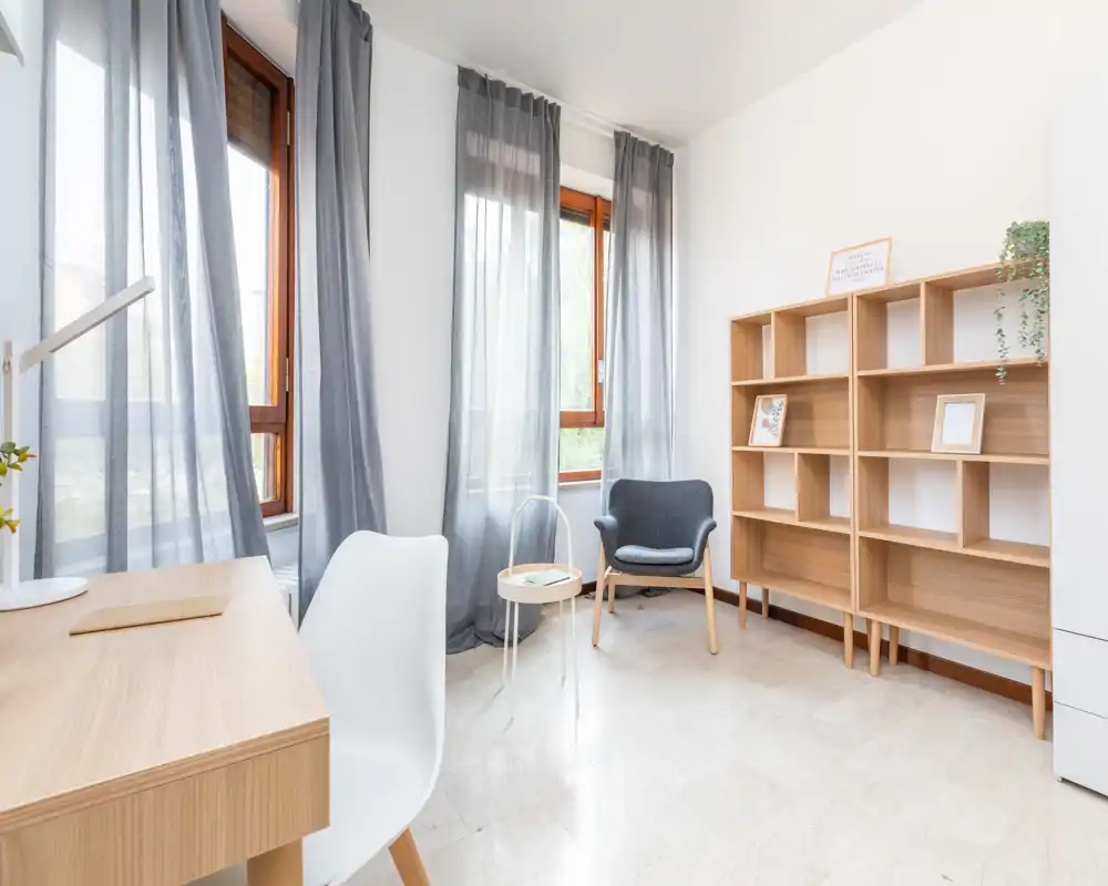 Zona de estar luminosa y minimalista con grandes ventanas, un sillón cómodo, estanterías y un pequeño escritorio — ideal para relajarse o trabajar de forma ligera.