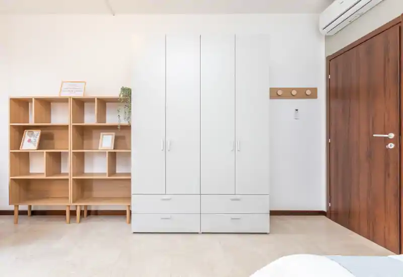 Minimalist bedroom corner with a white wardrobe, wooden shelving and a partial view of the bed; bright, clean styling ideal for listing photos.