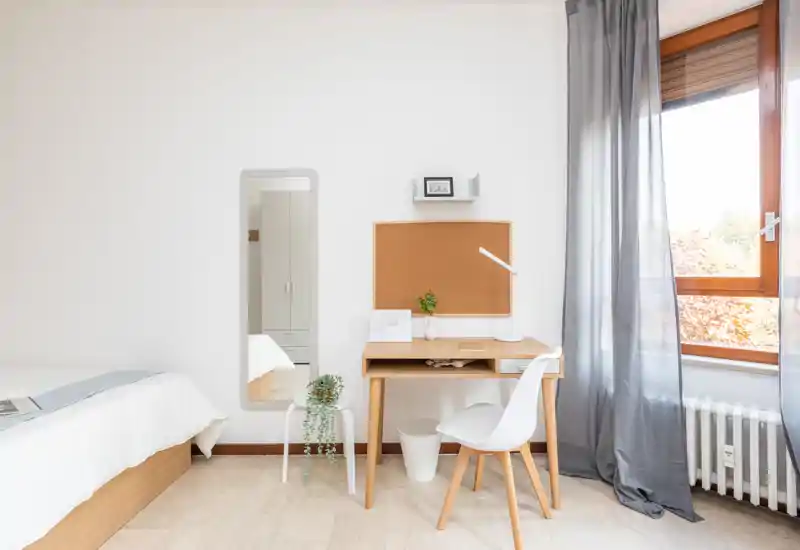 Dormitorio luminoso y minimalista con estructura de cama de madera, pequeño escritorio y luz natural desde la ventana. Paleta neutra y decoración sencilla que crean un ambiente tranquilo y ventilado.