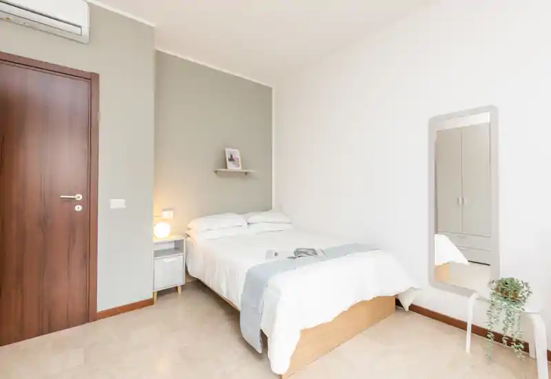 Dormitorio luminoso y minimalista con cama doble, mesita con lámpara, balda en la pared y espejo de cuerpo entero que refleja un armario. Tonos neutros, ropa de cama suave y mobiliario moderno y sencillo, perfecto para una estancia tranquila.