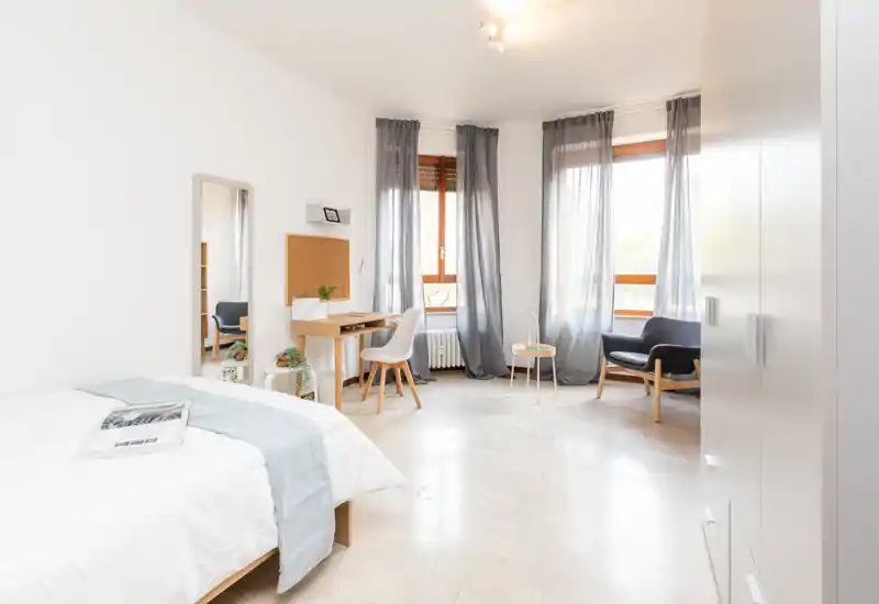 Dormitorio luminoso y amplio con cama doble cómoda, pequeño escritorio de madera, butaca y grandes ventanas con visillos — ideal para relajarse o trabajar.