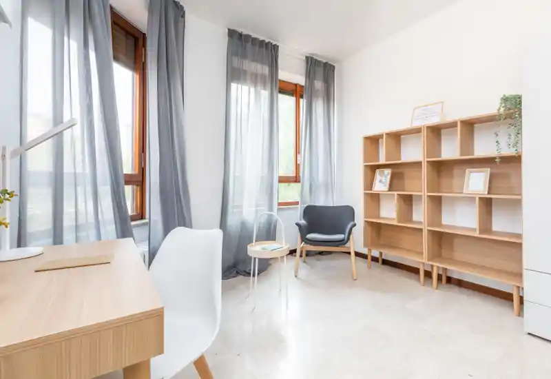 Bright, minimalist living area with large windows, a comfortable armchair, shelving and a small desk — ideal for relaxing or light work.