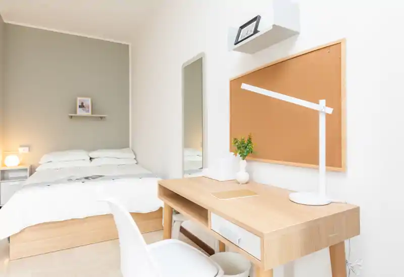 Dormitorio luminoso y minimalista con una cama doble cómoda, lámpara de noche y un escritorio de madera ordenado — ideal para descansar y trabajar de forma ligera.