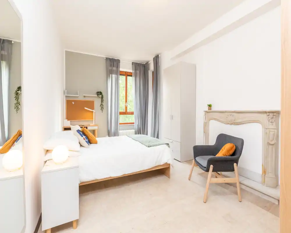 Camera da letto luminosa e moderna con letto matrimoniale ben rifatto, tessuti morbidi e una poltrona accogliente accanto a un camino decorativo. La luce naturale e i toni neutri creano un'atmosfera tranquilla e invitante, perfetta per gli annunci.