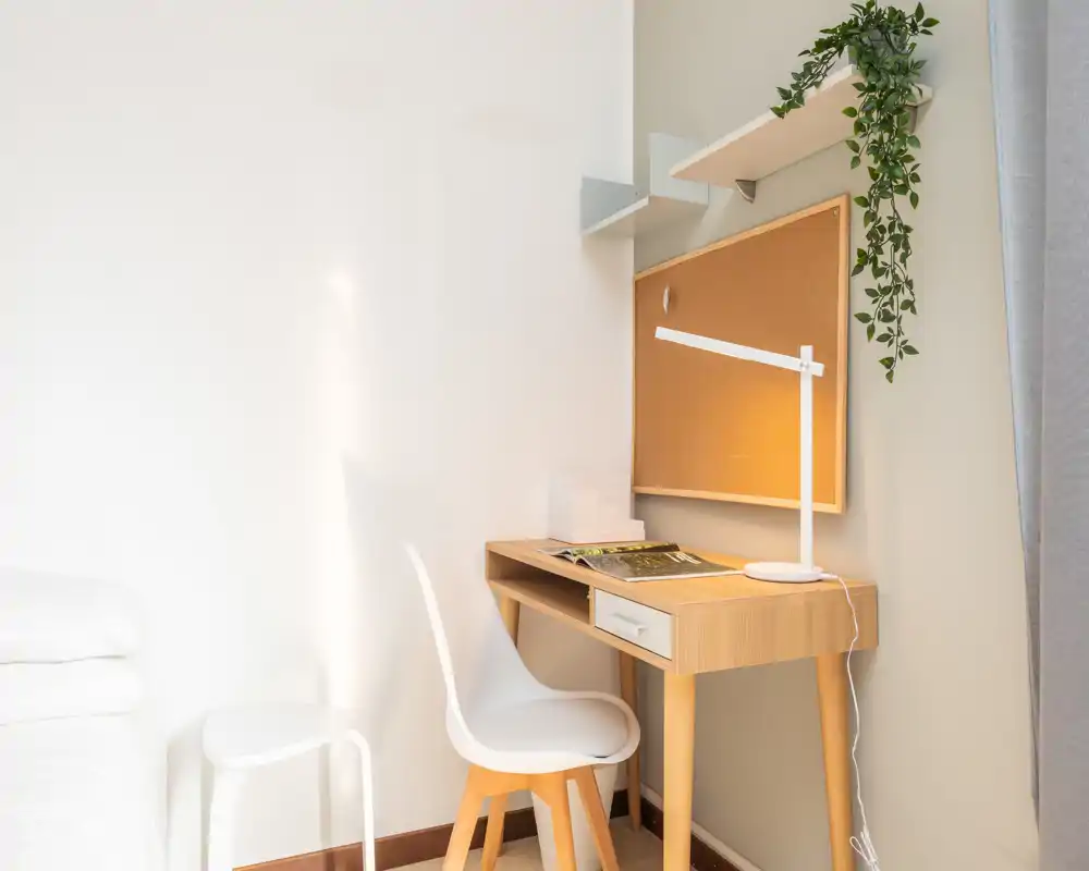 Angolo luminoso e minimale di una camera con scrivania in legno e sedia accanto a un letto parzialmente visibile. Stile pulito e moderno con bacheca e pianta: ideale come postazione di lavoro in una camera compatta.