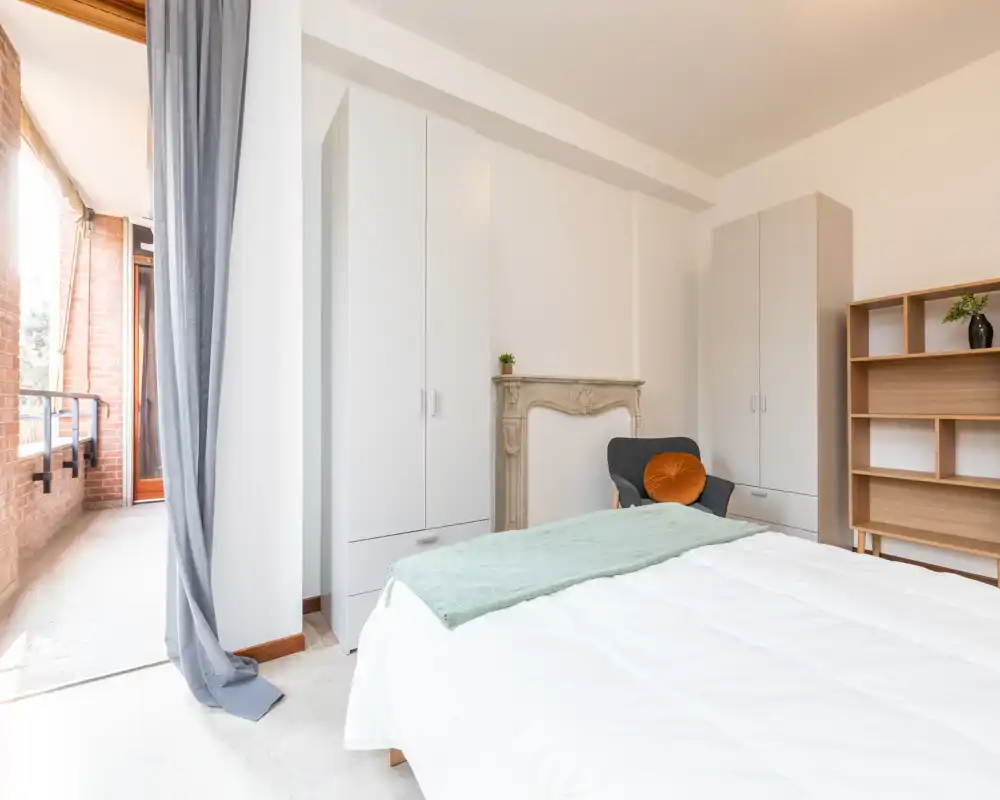 Camera da letto luminosa e minimale con letto ampio, armadi, una poltrona e accesso diretto al balcone che porta molta luce naturale.