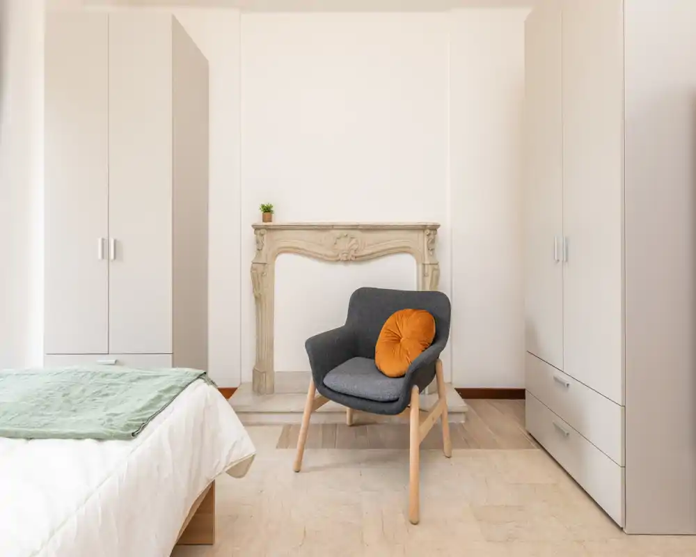 Camera da letto luminosa e minimale con letto parzialmente visibile, due armadi alti e una poltrona accogliente con cuscino arancione — linee pulite e toni neutri per un'atmosfera tranquilla.