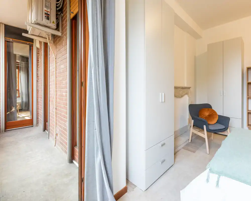 Camera da letto compatta e luminosa con letto visibile, armadio e una poltrona accogliente vicino al piccolo balcone; tessuti morbidi e toni neutri creano un’atmosfera rilassante.