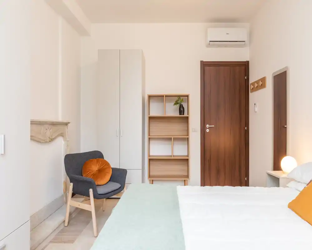 Camera da letto luminosa e moderna con letto ben rifatto, poltrona accogliente con cuscino decorativo, armadio e scaffale. Linee pulite e accenti caldi creano un ambiente invitante, perfetto per annunci.