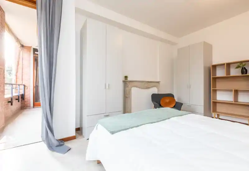 Dormitorio luminoso y minimalista con cama grande, armarios, un sillón acogedor y acceso directo al balcón que aporta luz natural.