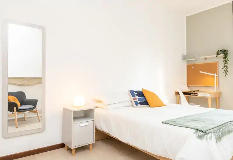 Dormitorio luminoso y minimalista con cama bien hecha, mesita de noche y pequeña zona de escritorio — ideal para una estancia tranquila con estilo moderno.