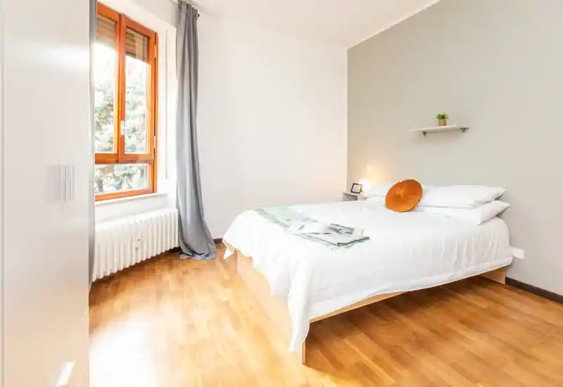 Camera da letto luminosa e minimal con letto matrimoniale, biancheria bianca e pavimento in legno caldo; luce naturale dalla finestra e dettagli decorativi morbidi la rendono accogliente.