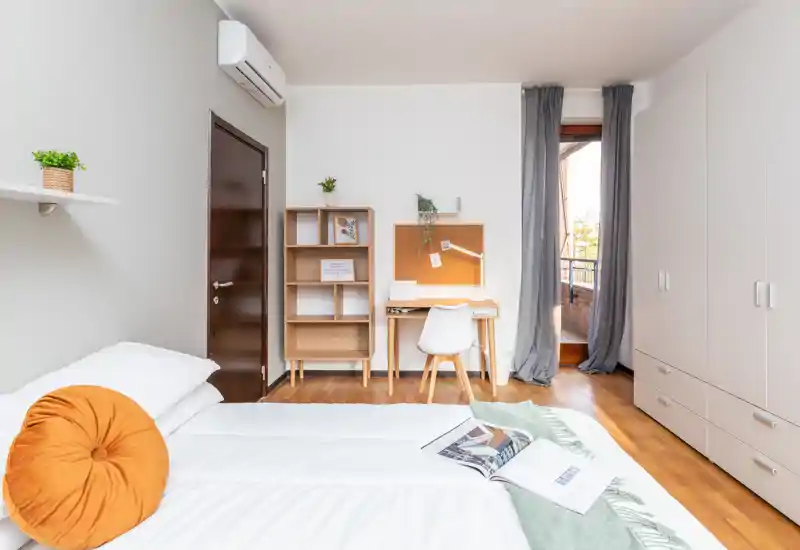 Camera da letto luminosa e moderna con letto accuratamente rifatto, cuscino rotondo decorativo e una scrivania vicino alla finestra con accesso al piccolo balcone.