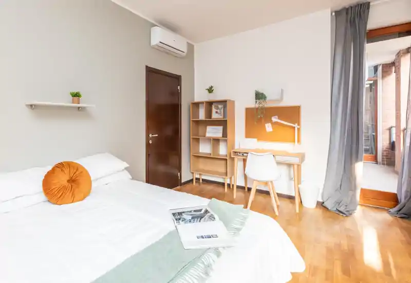 Camera da letto luminosa e moderna con un ampio letto in primo piano, una scrivania e una libreria vicino a una porta finestra che dà sul balcone. Biancheria bianca pulita, pavimento in legno caldo e dettagli decorativi rendono l'ambiente accogliente.