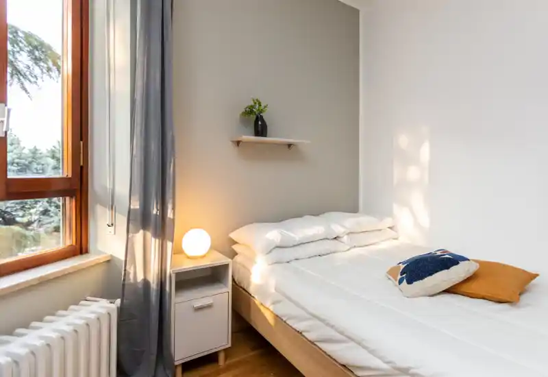 Camera da letto luminosa e minimal con letto rifatto, comodino con lampada e una grande finestra che lascia entrare luce naturale — perfetta per un soggiorno tranquillo.
