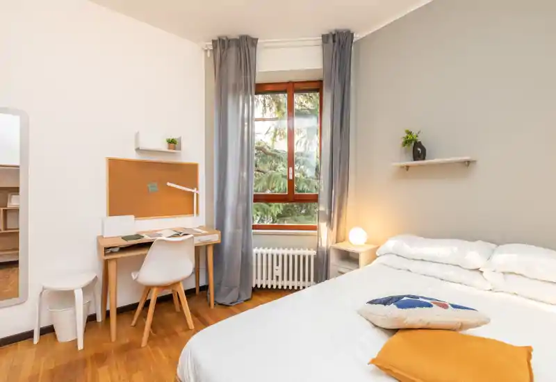 Camera da letto luminosa e moderna con un comodo letto matrimoniale, lampada da comodino e una piccola scrivania vicino alla finestra. Toni neutri, pavimento in legno e tessili morbidi creano un’atmosfera accogliente.