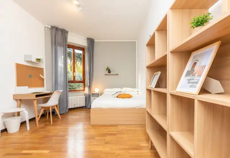 Camera da letto luminosa e ordinata con letto matrimoniale, lampada sul comodino e scrivania vicino alla finestra; pavimento in legno e scaffalature calde creano un ambiente accogliente e moderno.