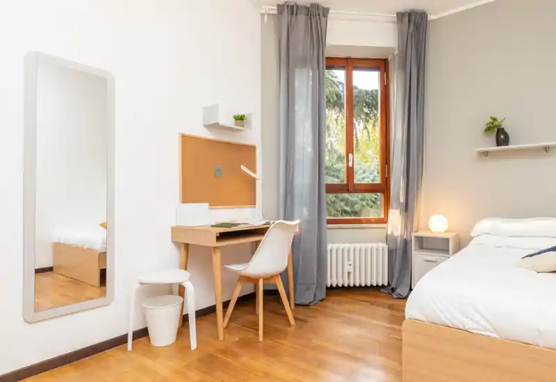 Camera da letto luminosa e ordinata con letto in legno visibile, comodino e una scrivania vicino alla finestra. Grande finestra con tende che offre luce naturale e atmosfera accogliente.