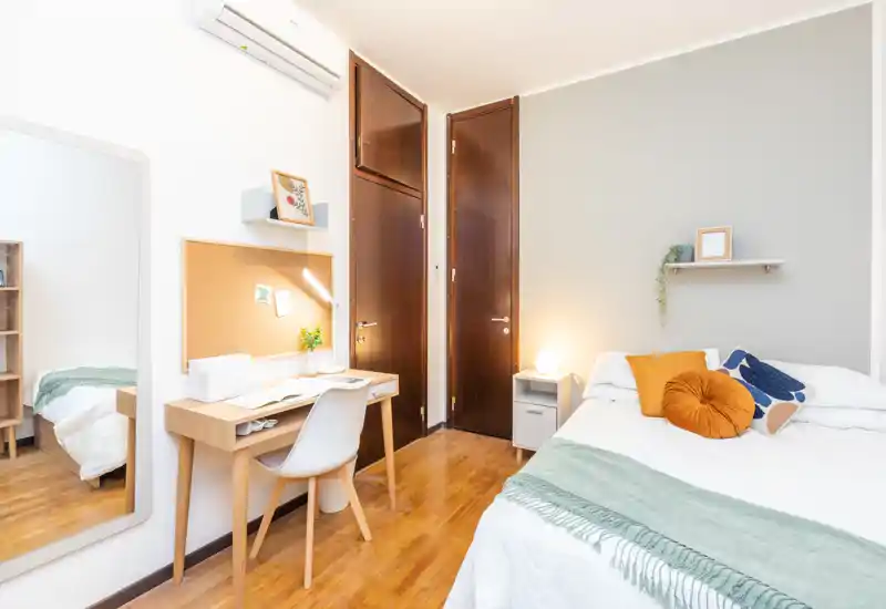 Camera da letto luminosa e ben arredata con letto matrimoniale, cuscini e plaid, un piccolo comodino con lampada e una scrivania compatta — ideale per studenti o professionisti.