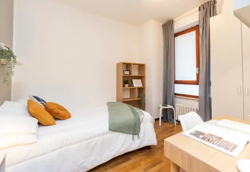 Camera da letto luminosa e ordinata con un letto ben visibile, tessili morbidi e una piccola zona studio vicino alla finestra. Pavimento in legno caldo, pareti neutre e cuscini decorativi per un ambiente accogliente.