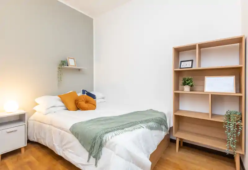 Camera da letto luminosa e accogliente con letto ben rifatto, cuscini decorativi e una libreria in legno — perfetta per foto di affitti.