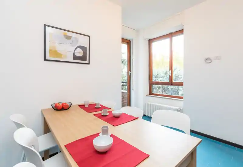 Bright, modern dining area with a wooden table set with red placemats and white chairs beside large windows letting in natural light.