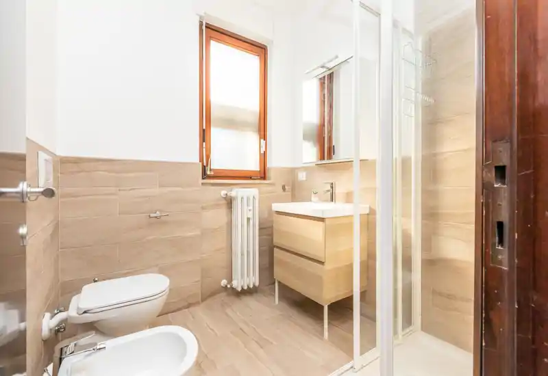 Bagno moderno e luminoso con wc a parete, bidet, piastrelle effetto legno, mobile lavabo e doccia con box in vetro. Linee pulite e toni naturali per un ambiente funzionale.