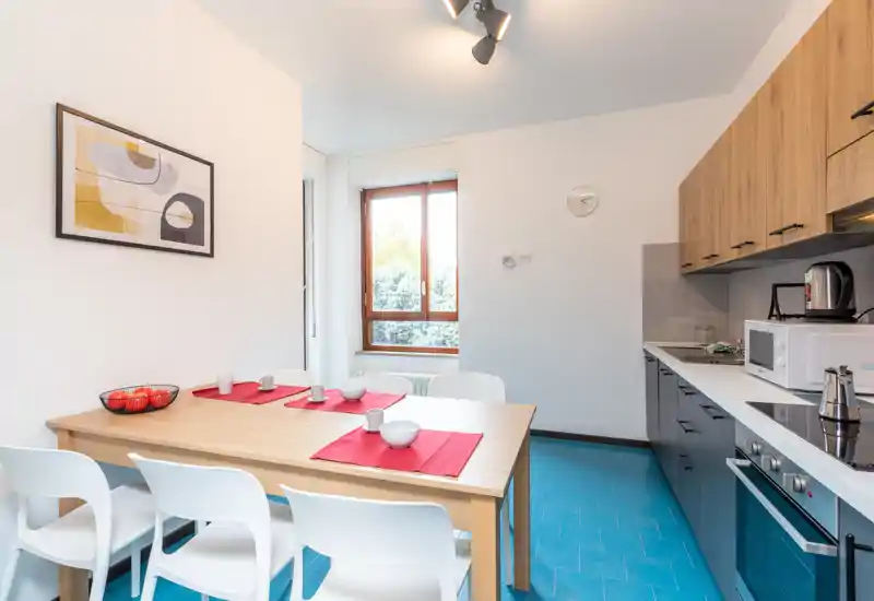 Modern kitchen with an integrated dining table: wood cabinets, built-in oven and microwave, and a wooden table set with red placemats and white chairs under bright natural light.