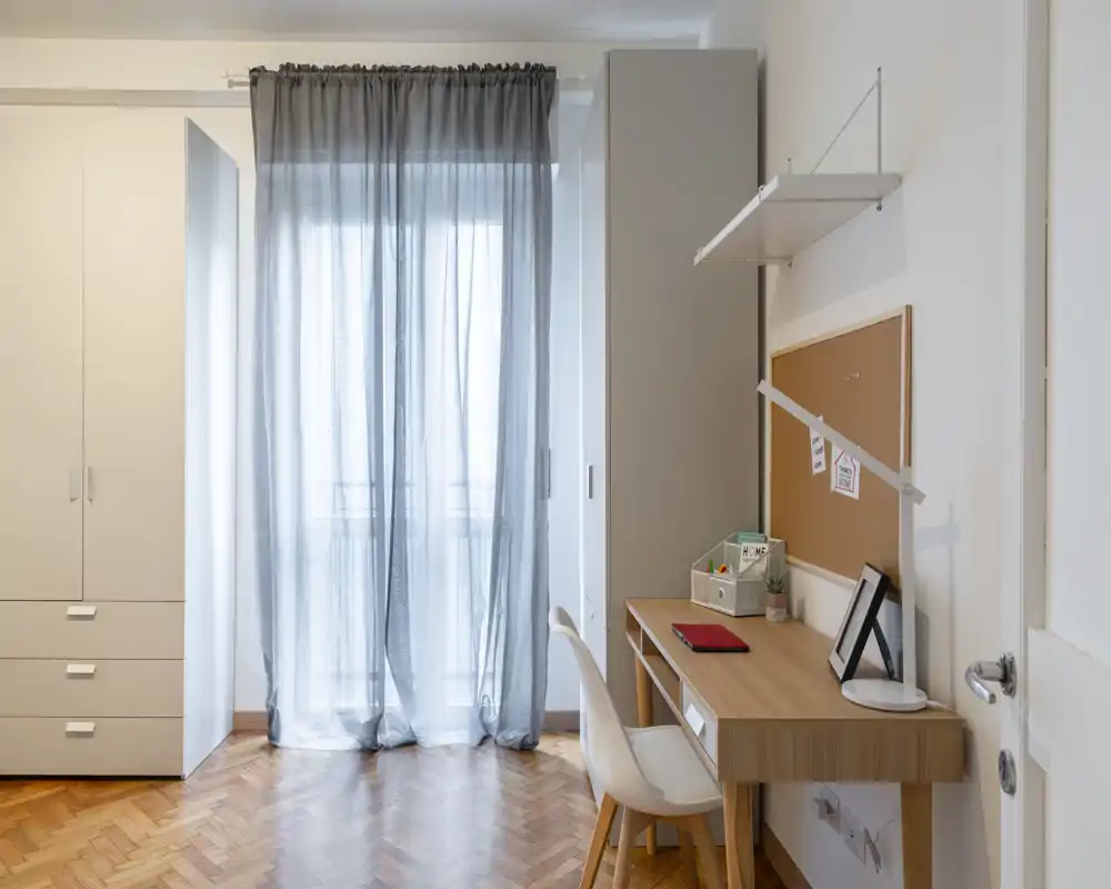 Angolo camera luminoso e ordinato con armadio a muro e scrivania in legno accanto a una finestra con tenda leggera; ideale per chi cerca uno spazio di studio tranquillo.