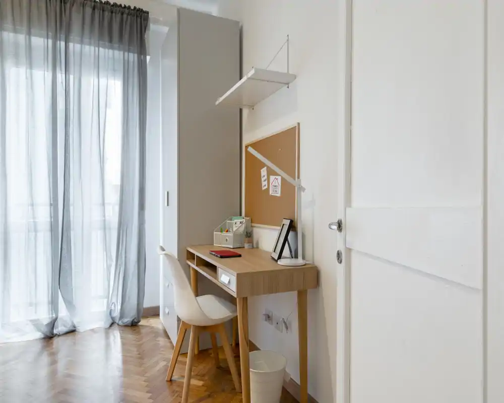Angolo studio luminoso e minimale con scrivania in legno, sedia bianca e bacheca vicino a una finestra con tende leggere — perfetto per uno spazio di lavoro compatto.