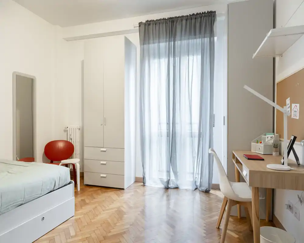 Camera da letto luminosa e minimale con letto singolo, armadio e scrivania vicino a una grande finestra che lascia entrare luce naturale.