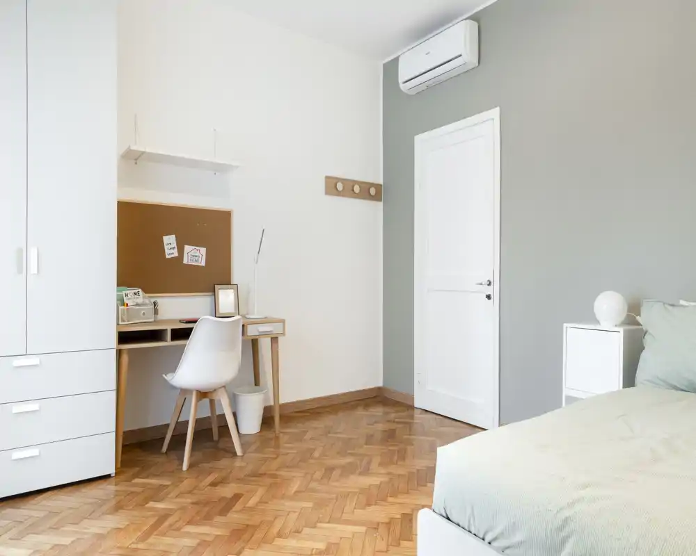 Camera da letto luminosa e minimal con letto sul lato destro, un comodino e una scrivania compatta. Pavimento in parquet e toni neutri creano un’atmosfera moderna e rilassante.