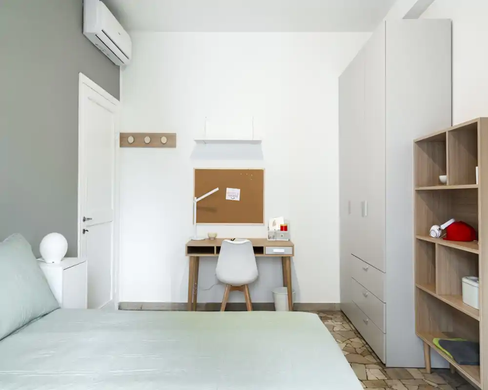 Camera da letto luminosa e minimal: letto ben visibile in primo piano, scrivania compatta con sedia sulla parete, grande armadio e scaffalatura aperta. Linee pulite e biancheria pastello creano un ambiente tranquillo e funzionale.