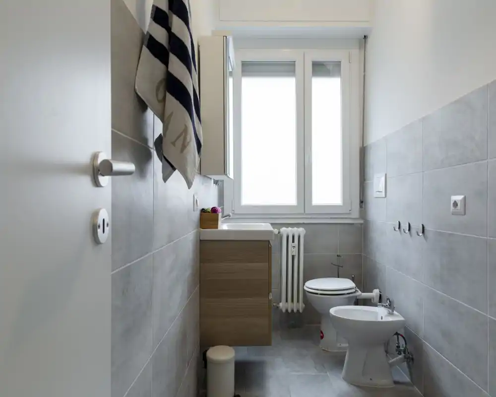 Bagno compatto e moderno con piastrelle, mobile lavabo sospeso, bidet e WC accanto a una finestra luminosa.