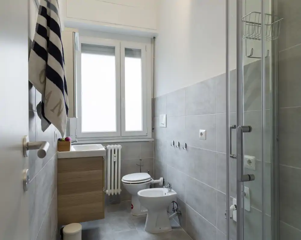 Bagno moderno e ben illuminato con box doccia, mobile lavabo, wc e bidet. Piastrelle grigie neutre e una grande finestra smerigliata assicurano un aspetto pulito e arioso.