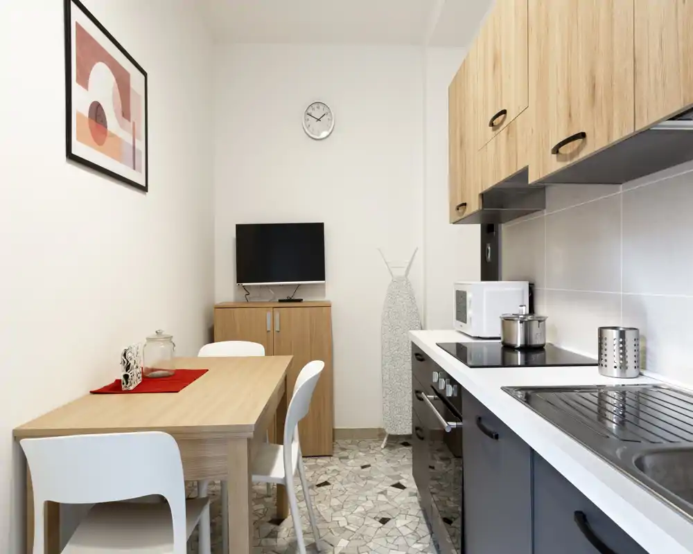 Cucina moderna compatta con piccolo tavolo da pranzo in legno, piano cottura integrato, microonde e lavello — luminosa, ordinata e funzionale per soggiorni brevi.