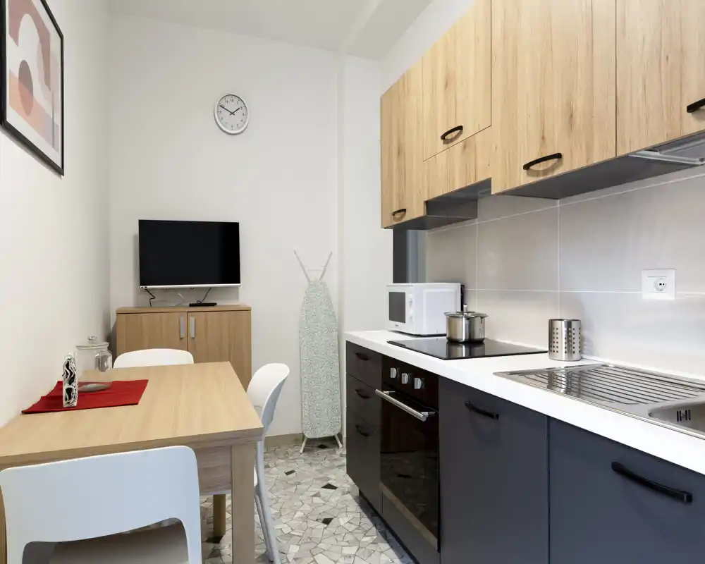 Cucina moderna compatta con forno integrato, piano cottura, microonde e lavello; un piccolo tavolo con sedie crea una pratica zona pranzo. Toni neutri, pensili in legno e pavimento piastrellato donano un aspetto pulito e contemporaneo, ideale per affitti brevi.