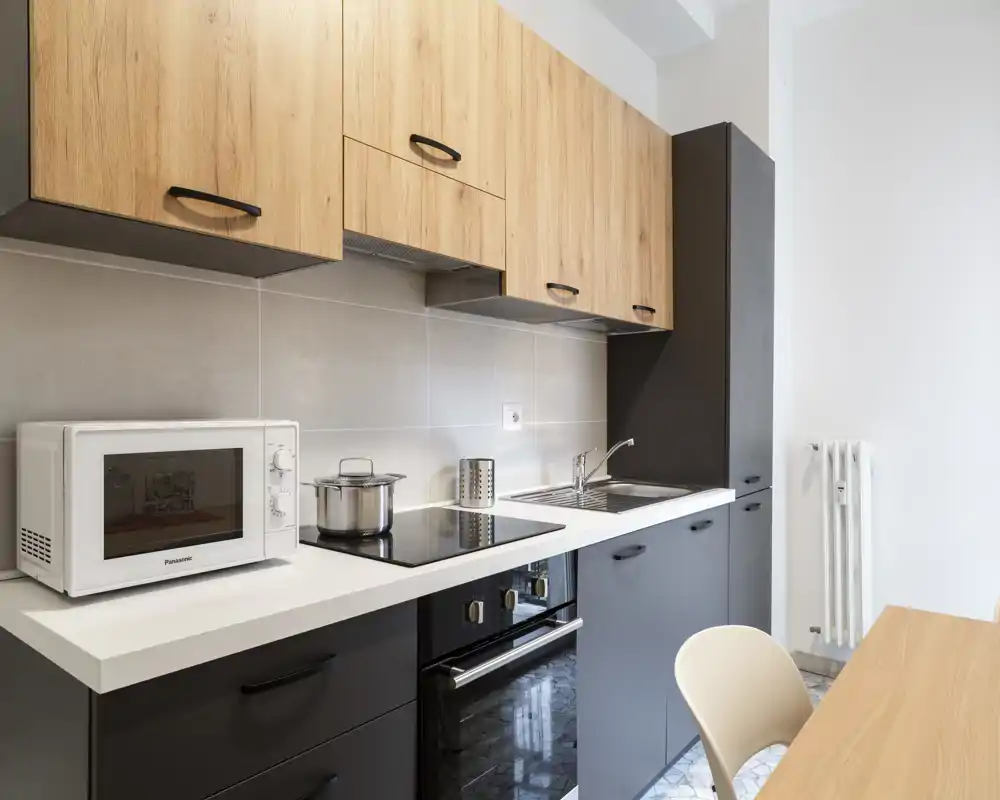 Cucina moderna e compatta con pensili in legno, basi scure, piano cottura a induzione, forno integrato, lavello e microonde sul piano. Spazio pulito e ben illuminato ideale per affitti brevi o monolocali.