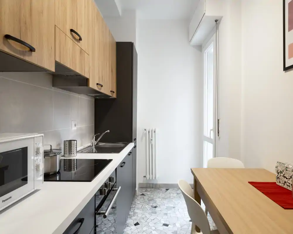 Cucina moderna a corridoio, luminosa, con mobili in legno, piano a induzione, microonde e piccolo tavolo da pranzo — ambiente pulito e funzionale con luce naturale.