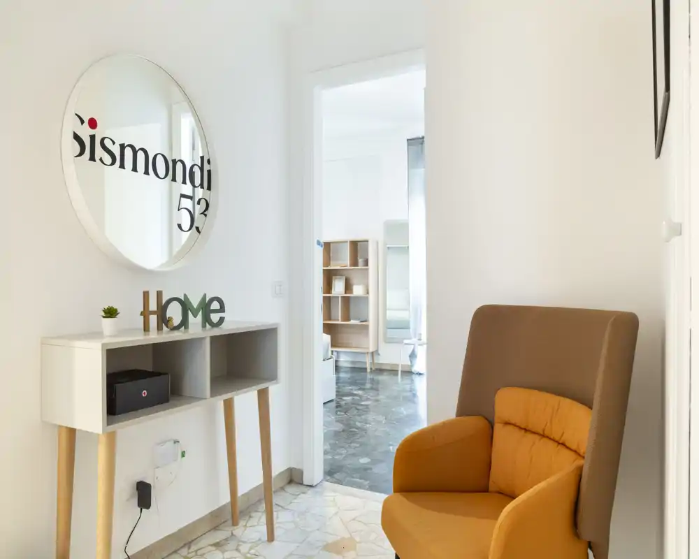 Ingresso luminoso e minimal con consolle, specchio rotondo e poltrona arancione; pavimento chiaro in piastrelle conduce a una stanza interna.