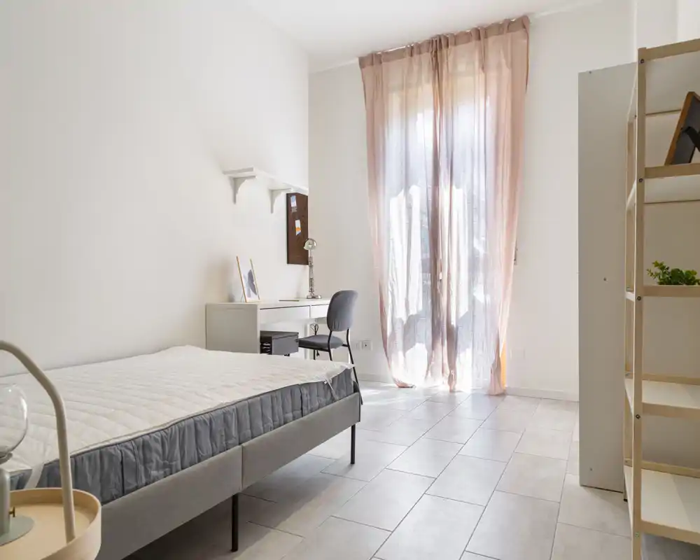 Bright, minimal bedroom with a single bed, desk and soft natural light from a large window with sheer curtains — ideal for a student or single occupant.