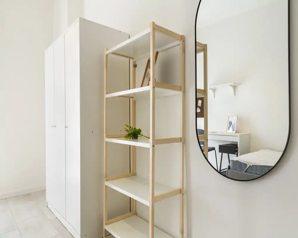 Minimalist bedroom corner with a white wardrobe, open shelving and a large wall mirror reflecting the bed and desk; bright, clean and airy space suitable for rental photos.