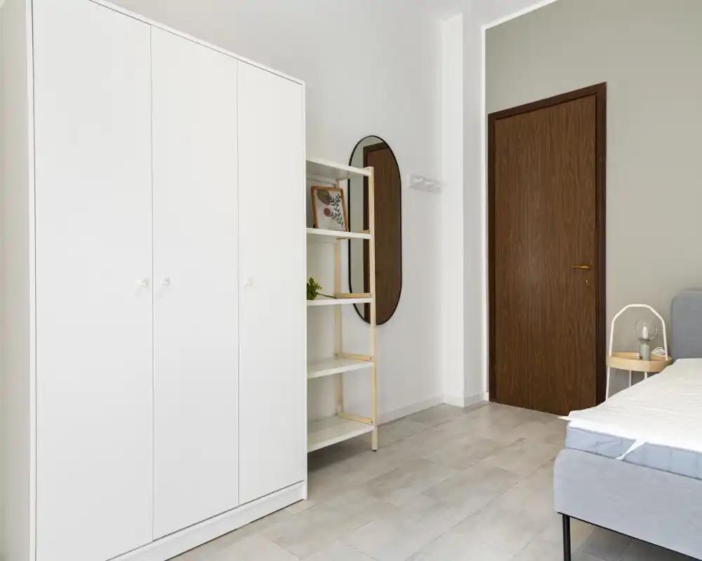 Minimalist bedroom corner with a grey upholstered bed, white wardrobe and shelving, wooden door and oval wall mirror; bright, clean and simply styled for rental listings.